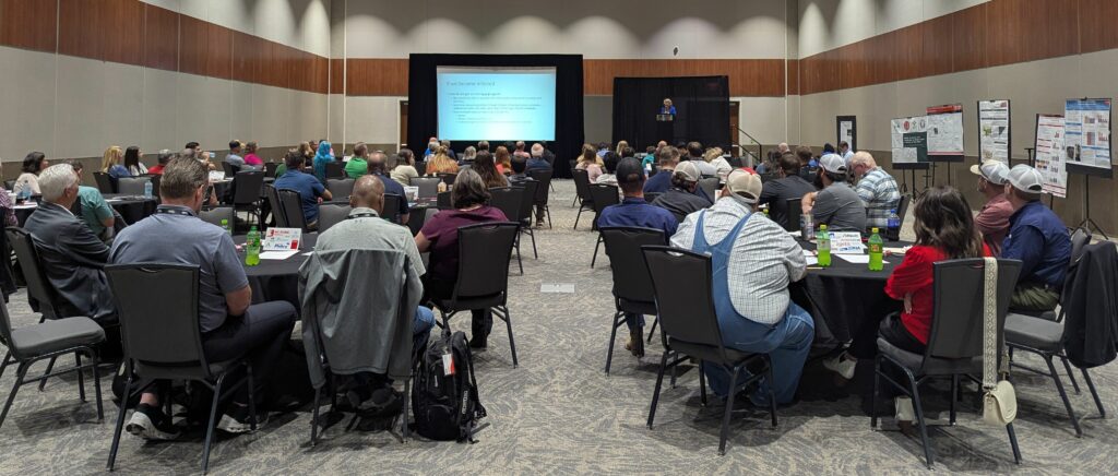 Conference attendees sit at round tables facing a speaker at the front of the room, with a presentation projected on a screen and educational posters displayed along the side wall.